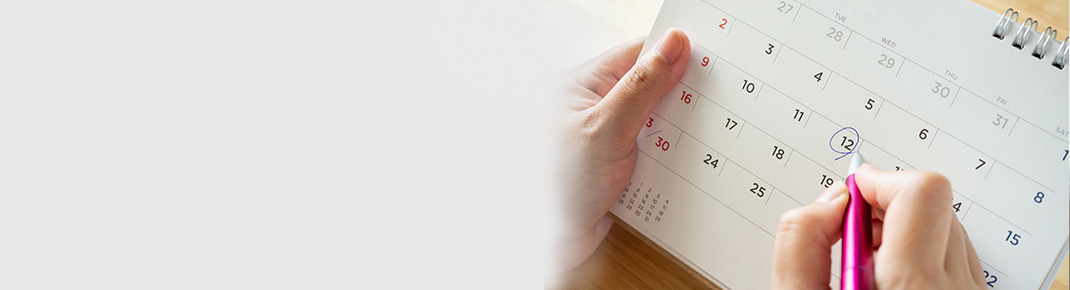 Kvinna i glasögon som bär en mörkblå skjorta lutar sig på en vit säng med ett leende som ser eftertänksamt. / Närbild på en kalender med en kvinnas hand som markerar en dag med penna.